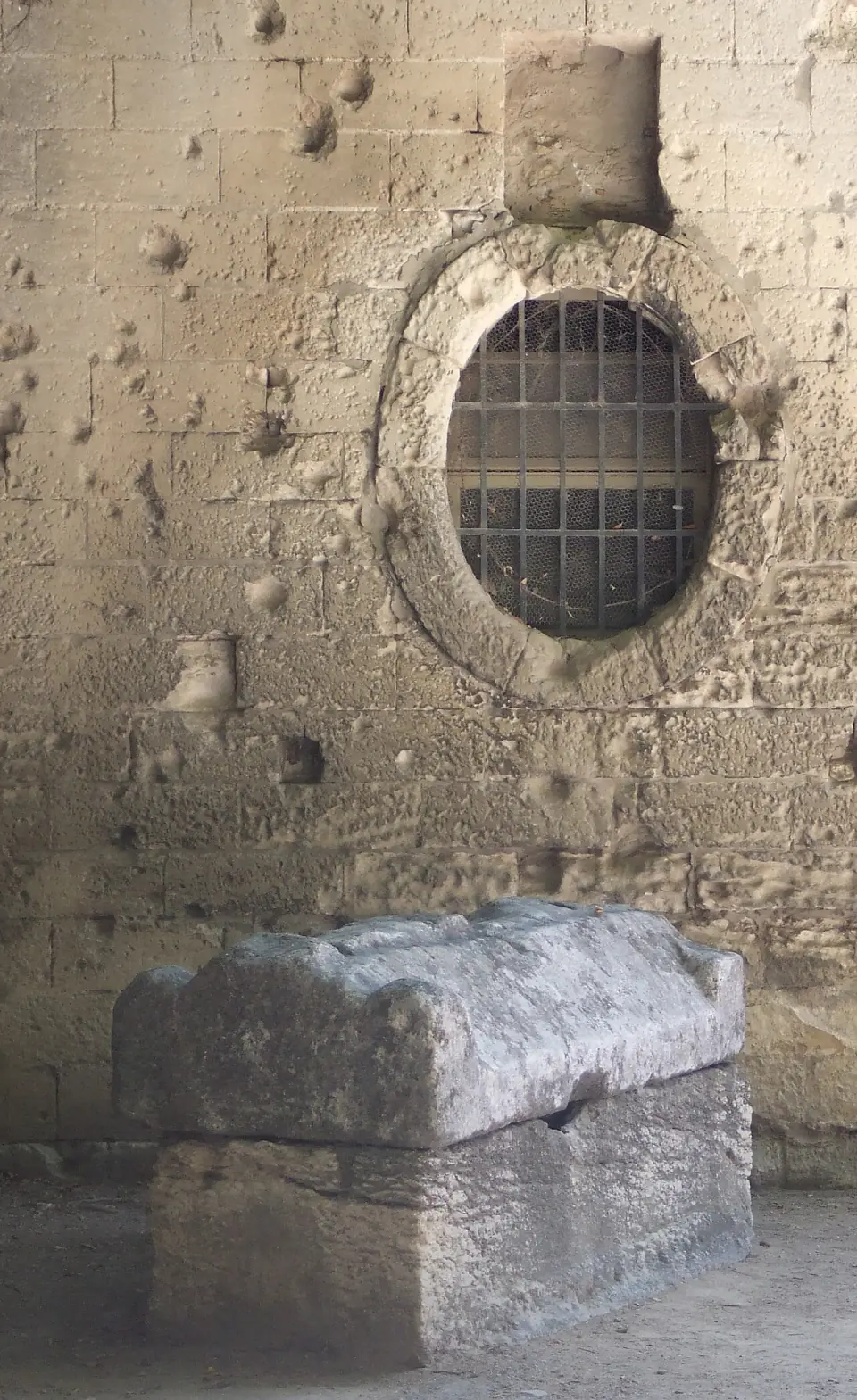 Un sarcophage de pierre posé sous un mur, devant une fenêtre ovale grillagée