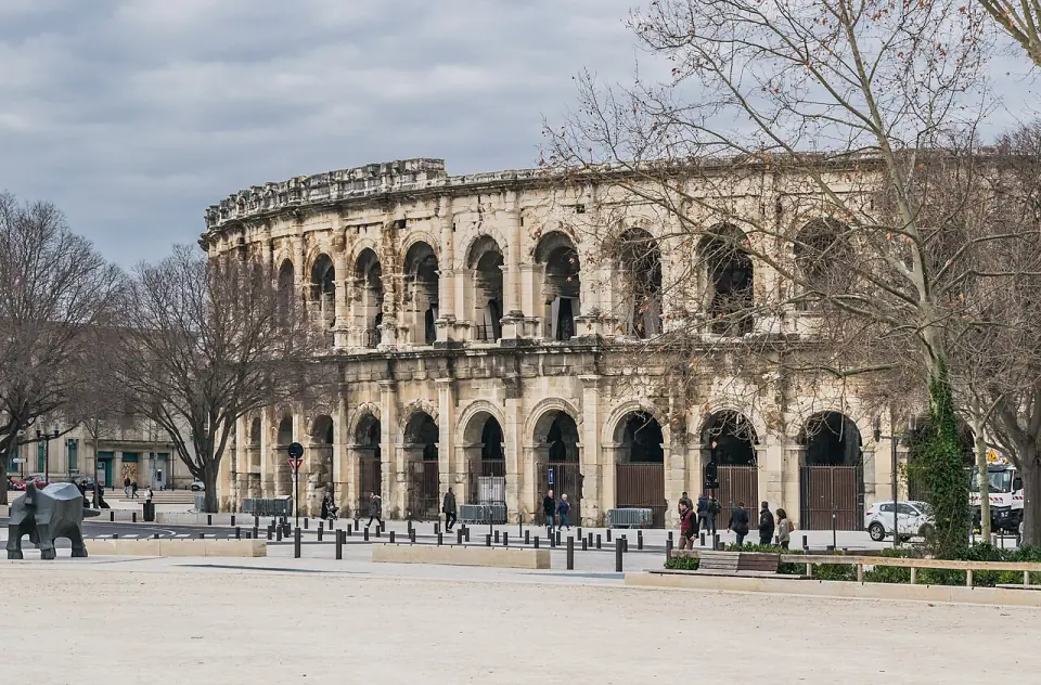 Les arènes de Nîmes vues depuis le parvis, derrière des arbres, sous un ciel nuageux