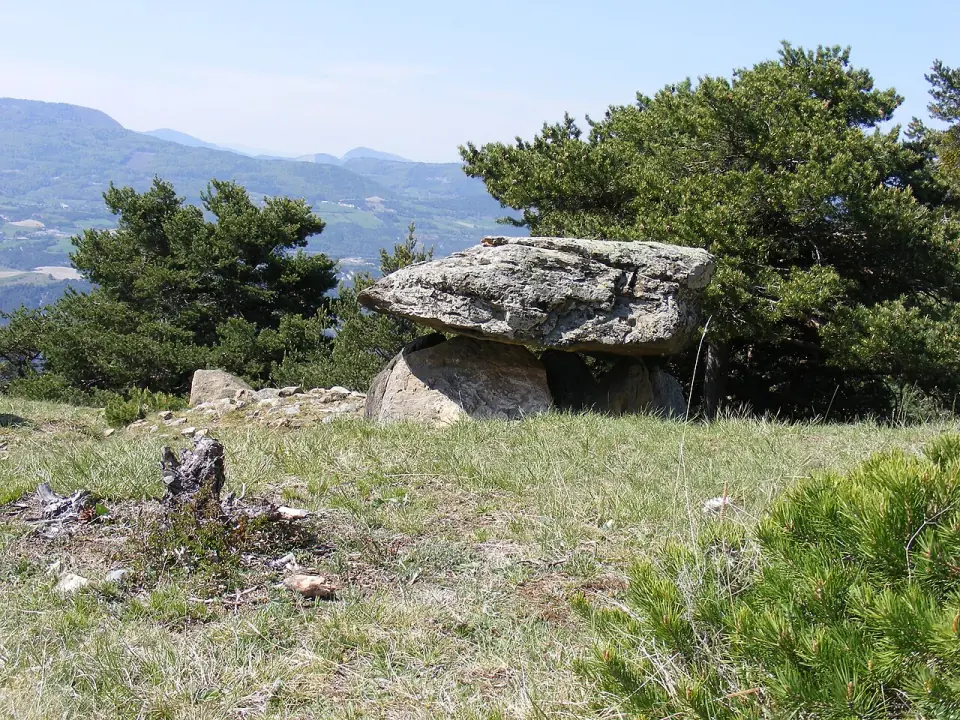 Le dolmen du Villard : grande dalle de couverture posée sur des pierres, au milieu des pins, avec des montagnes au loin