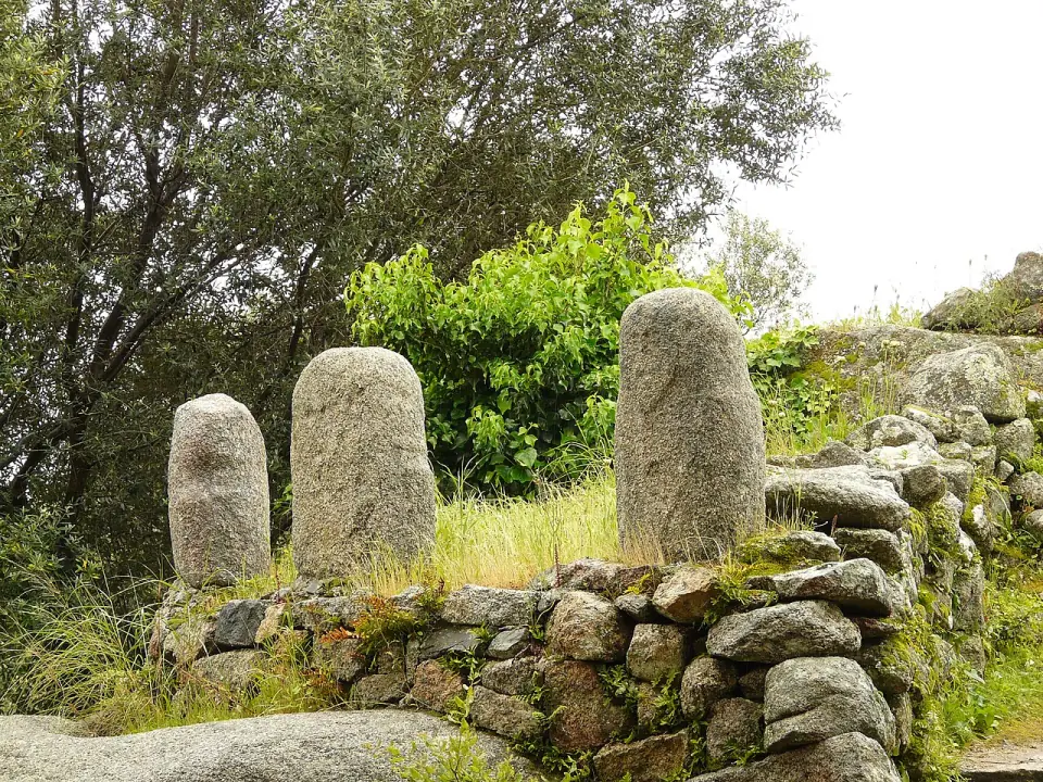 Trois statues-menhirs en granite alignées au-dessus d'un mur de pierres sèches, parmi la végétation