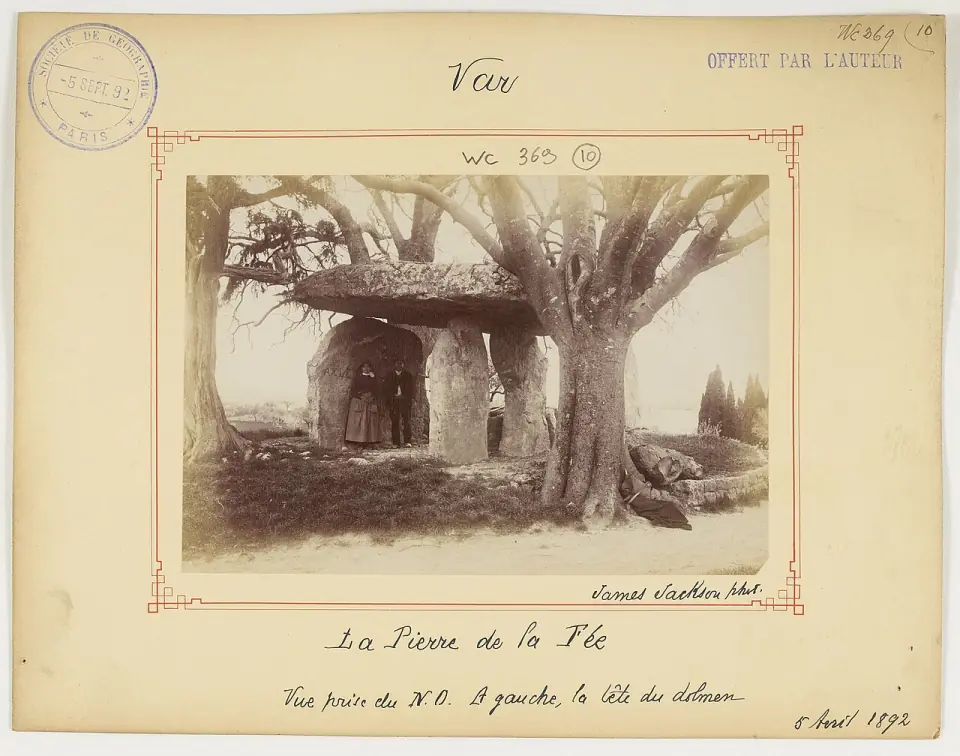 Photographie de 1892 de la Pierre de la Fée entre deux grands arbres, avec deux personnes sous la dalle