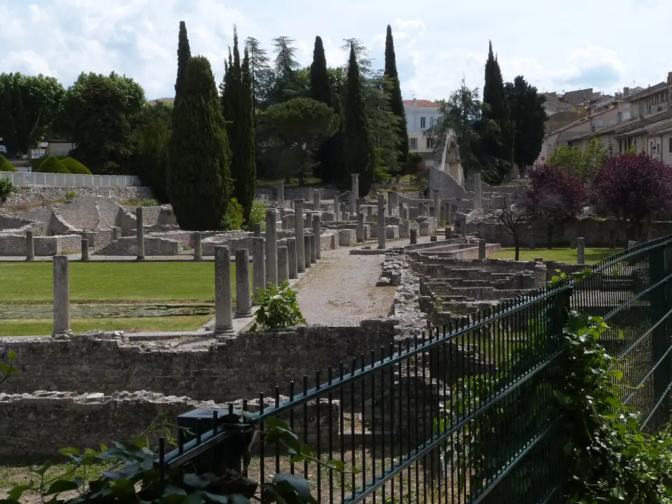 Vue du champ de fouilles de la Villasse derrière une grille, avec cyprès et colonnes